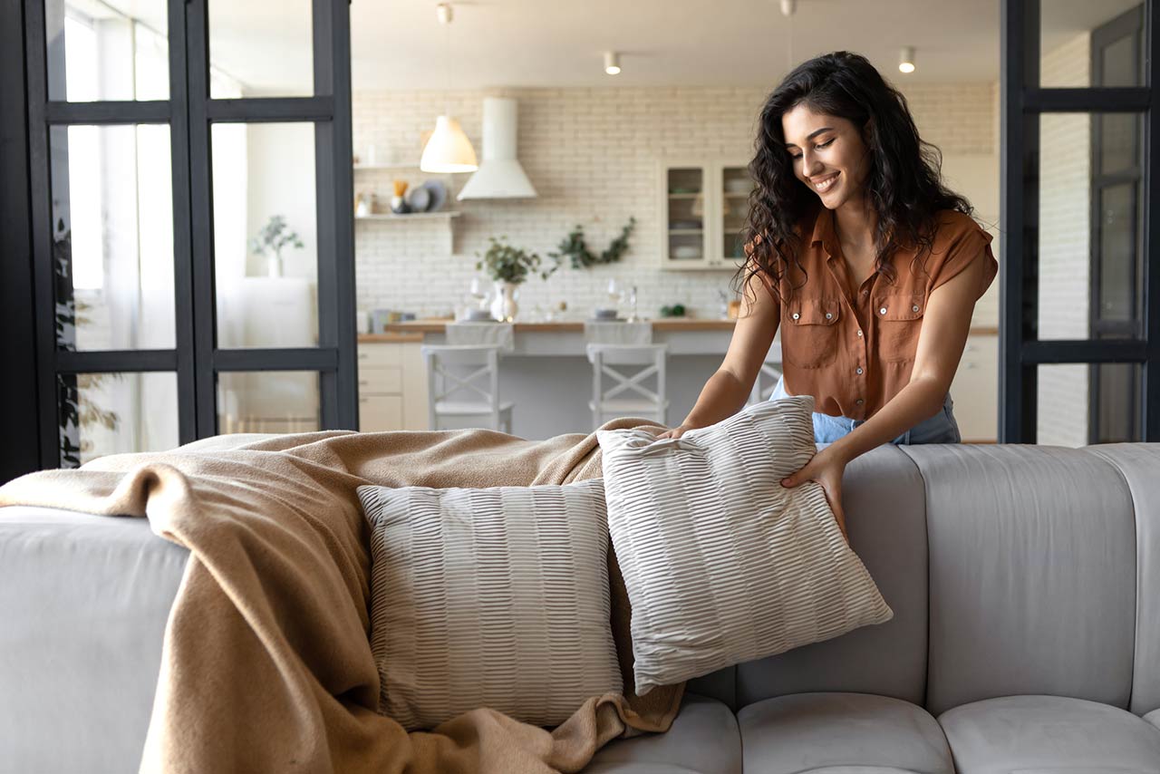 Lovely young woman putting soft pillows and plaid on comfy sofa, making her home cozy and warm